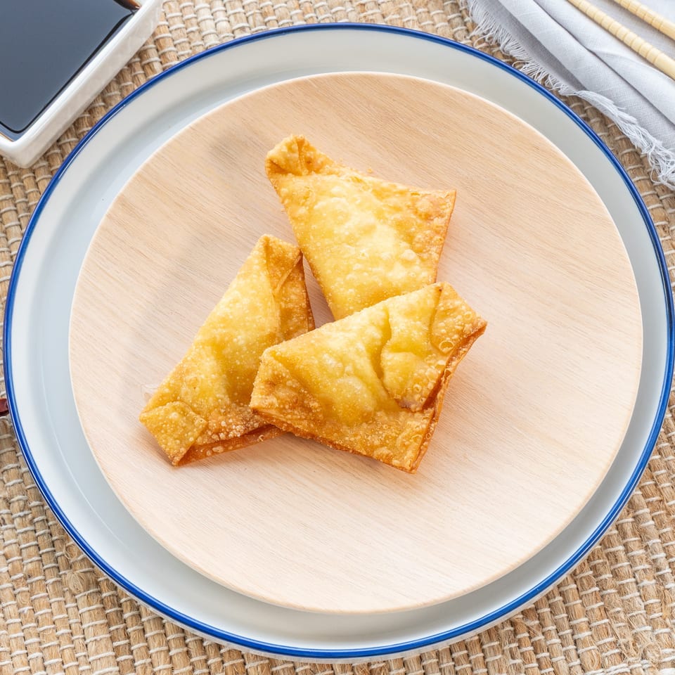 Three crispy, golden-brown crab rangoons on a light wooden plate, with soy sauce and chopsticks nearby.