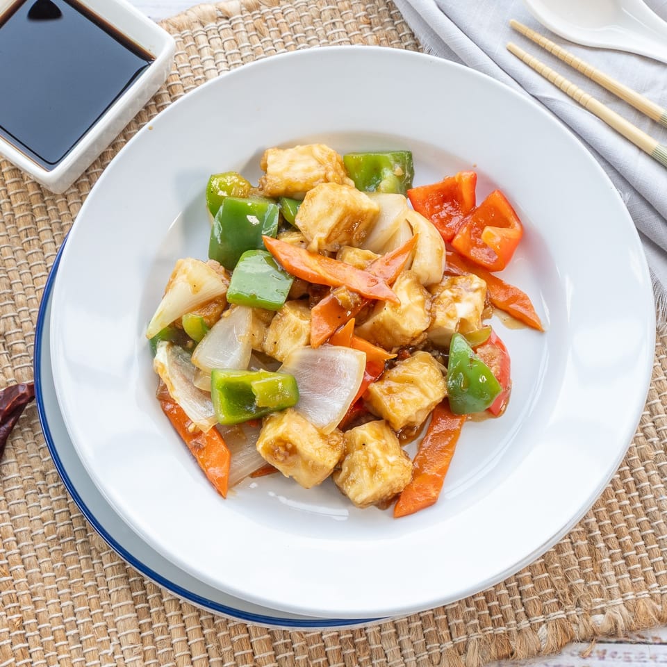 Sweet and sour tofu with bell peppers, onions, and carrots on a white plate with soy sauce.