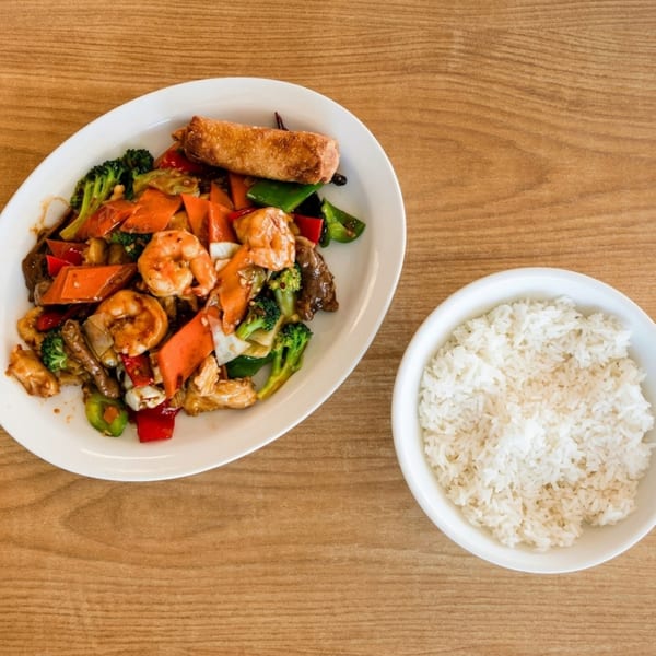 Shrimp and vegetable stir-fry with an egg roll and a side of white rice on a wooden table.