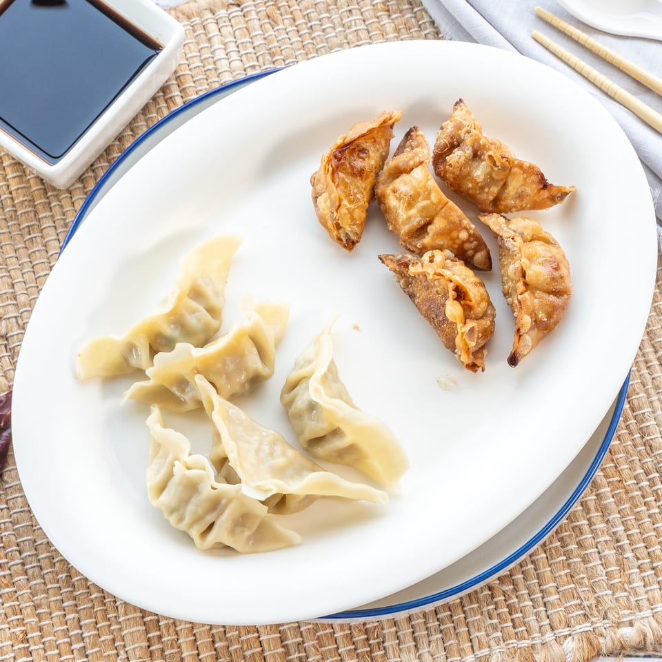 A white oval plate holds steamed and fried Chinese dumplings, with soy sauce and chopsticks nearby.
