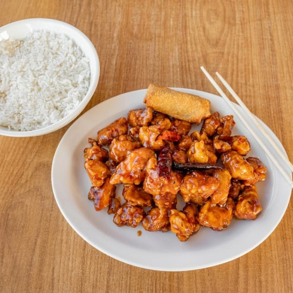 A plate of orange chicken with an egg roll and chopsticks, next to a bowl of white rice.